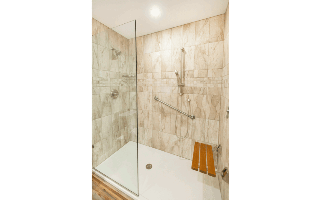 Frameless glass shower door on remodeled standing shower with white marble-look wall tiles, a wooden seating bench to the right, and grab bars on the wall.