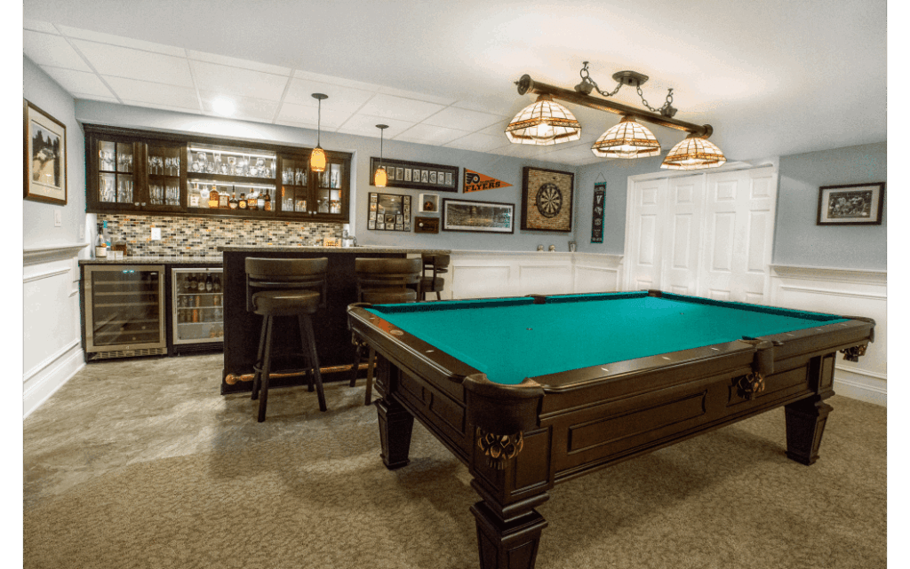 A finished basement with light gray/tan flooring, a pool table in foreground, and a small bar area with dark wood cabinets in background.