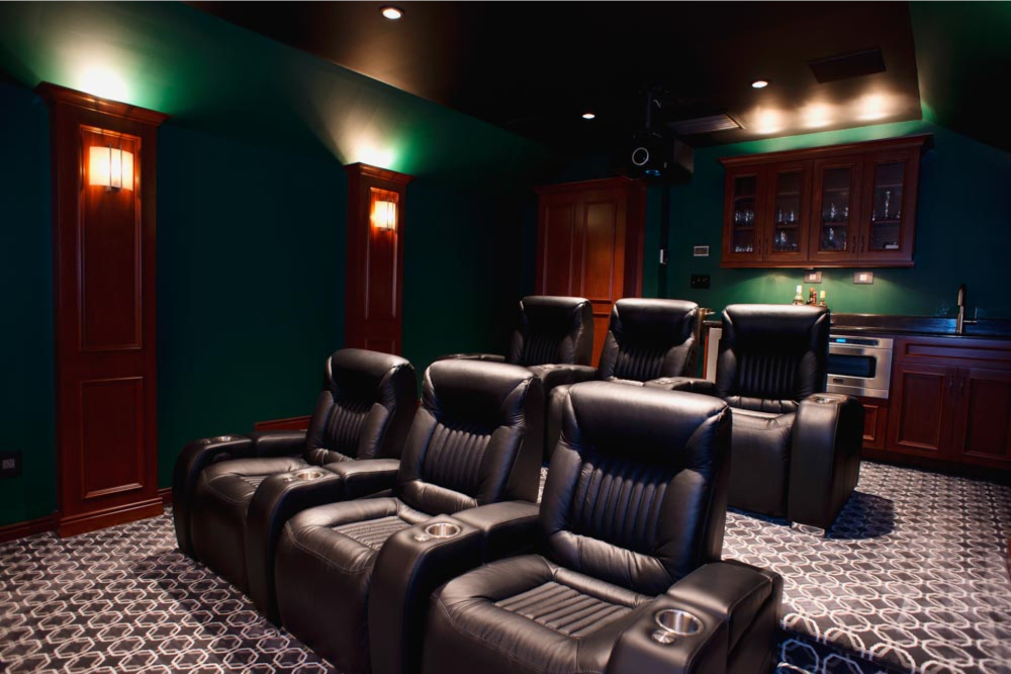 Newly added home theater with two rows of three leather recliners each, with cupholders, dark cabinets for small bar area along back wall. Dark green walls with sconce lighting, and carpeted floor.