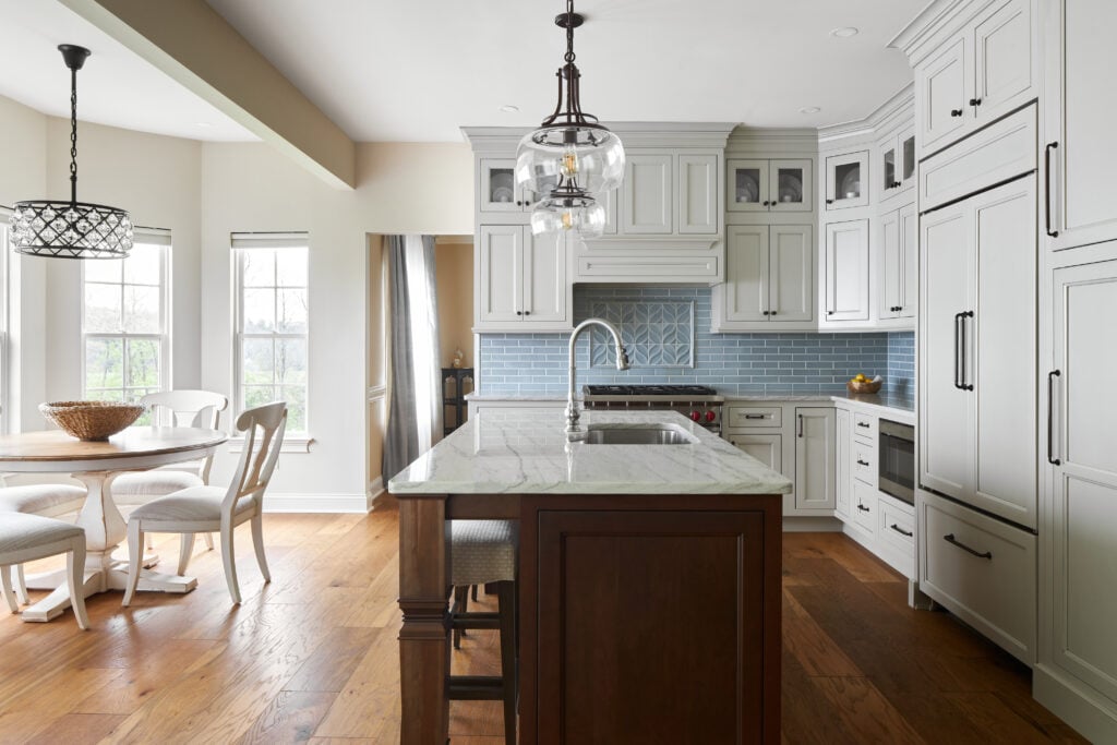 Wide-plank mid-tone engineered hardwood in a remodeled white kitchen with woodgrain island, white stone countertops, light blue tile backsplash, and wood-and-white dining table in dining area to the left.