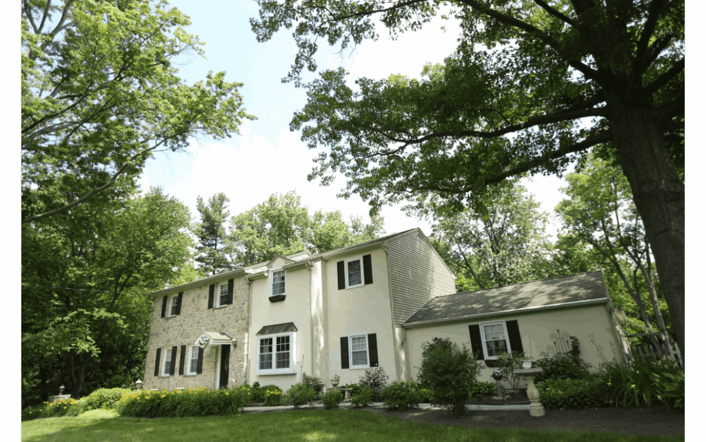 Exterior view of large colonial style home with light brick and pale yellow stucco siding and black shutter, with a one-story addition on the right side. Trees surrounding.