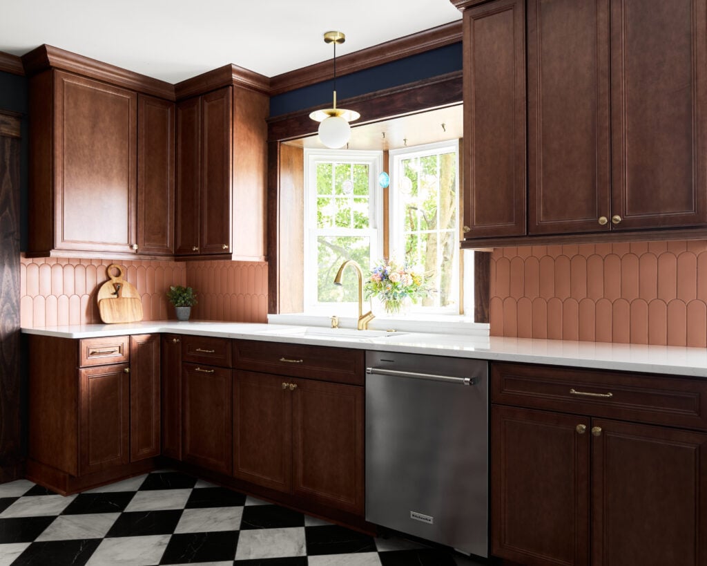 Woodgrain transitional style cabinets in a home with gold hardware, black-and-white tiled floor, and retro peach glass tile backsplash. White stone countertops.