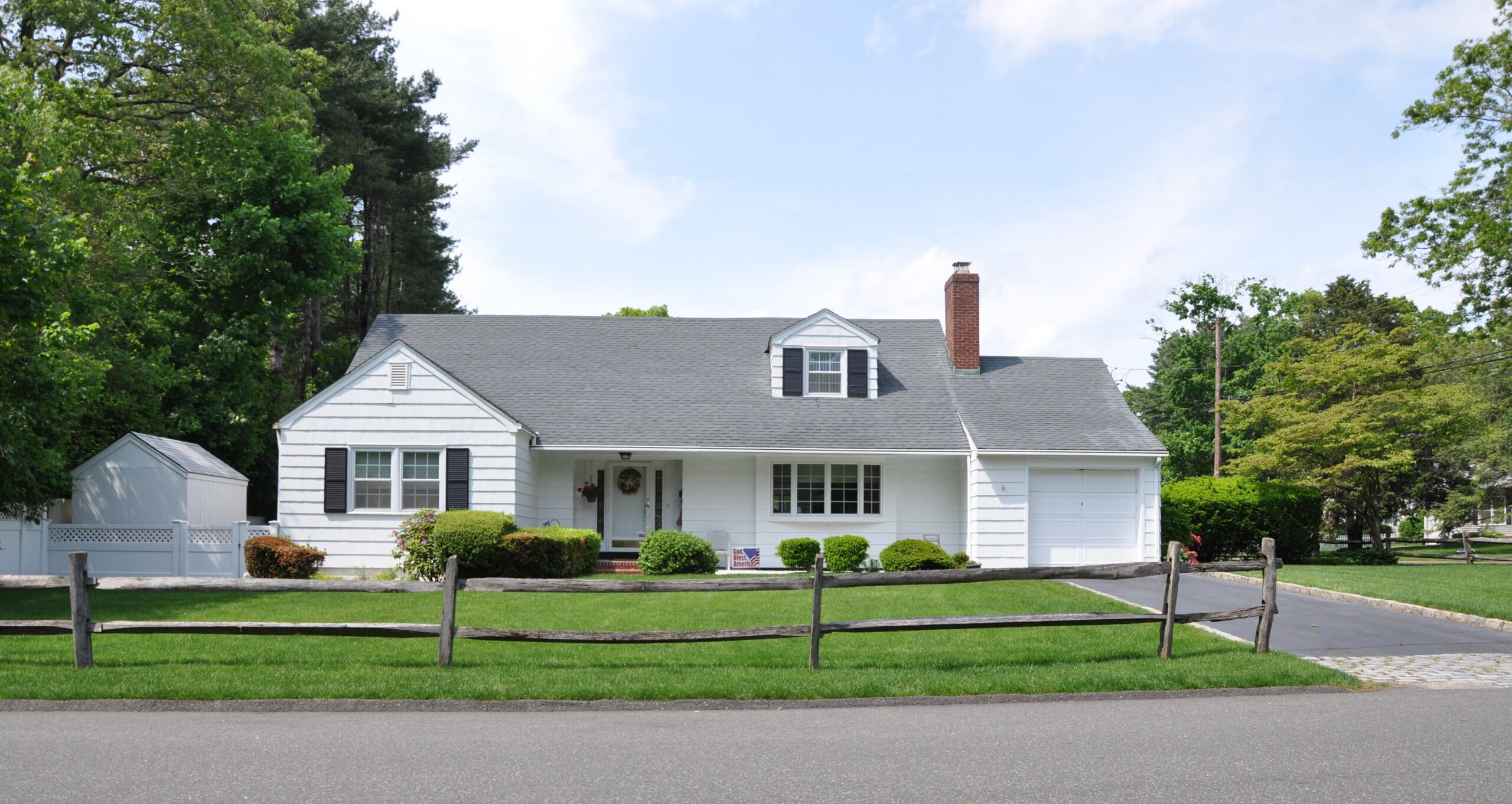 White ranch style home with black shutters, manicured green front lawn, one-car garage on right, and trees on either side. Dormer addition.