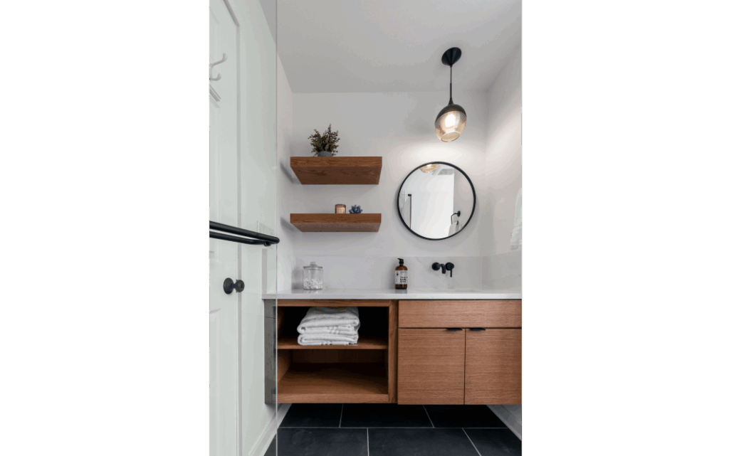 Remodeled woodgrain bathroom vanity with storage underneath and two floating wood plan shelves on the wall, next to a round mirror.