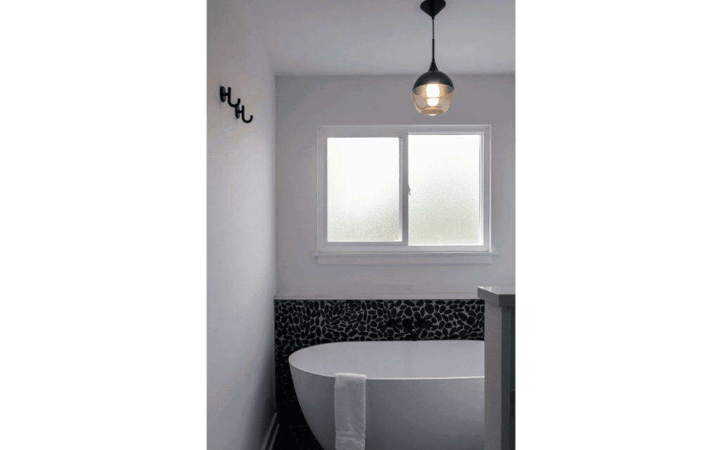 Large soaking tub sitting under foggy glass window, with statement black pebble-tile on the bottom half of the wall. Black pendant light over the tub.