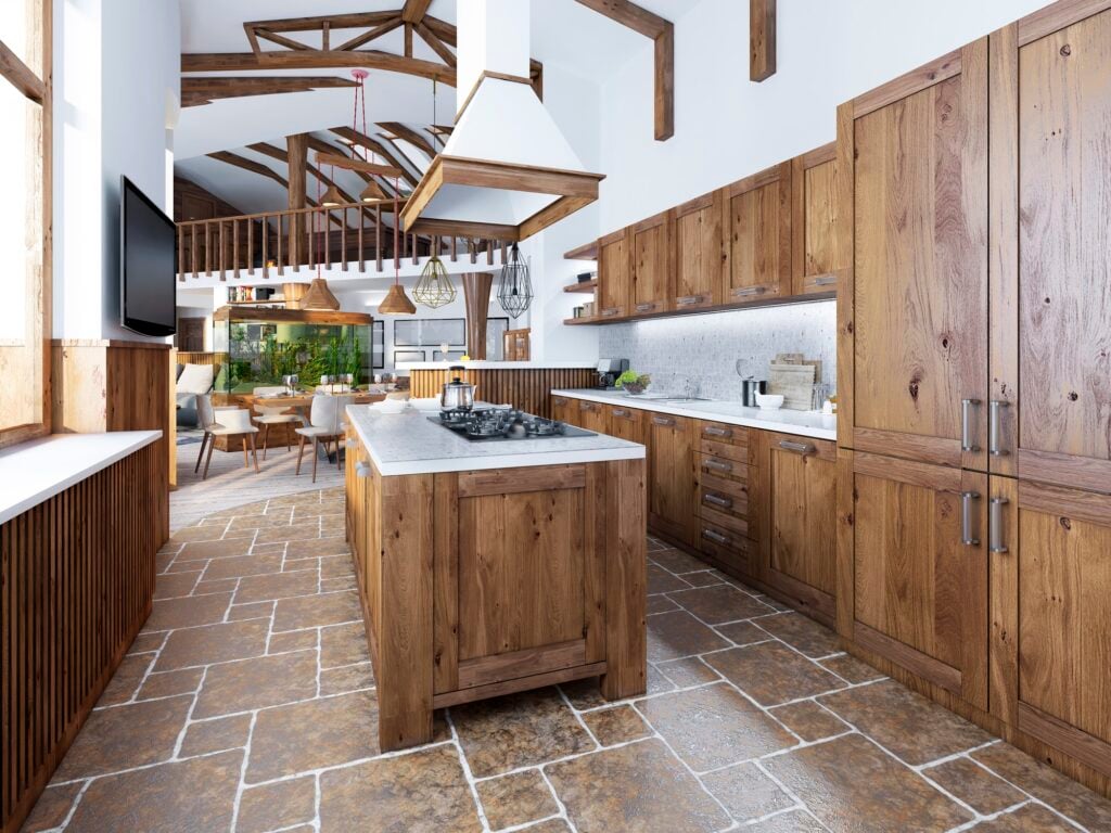 Large kitchen with an island in the middle. Wooden furniture with white countertops and mosaic with integrated appliances. Brown tile floor. Woodgrain cabinets. Wood beams on ceiling.
