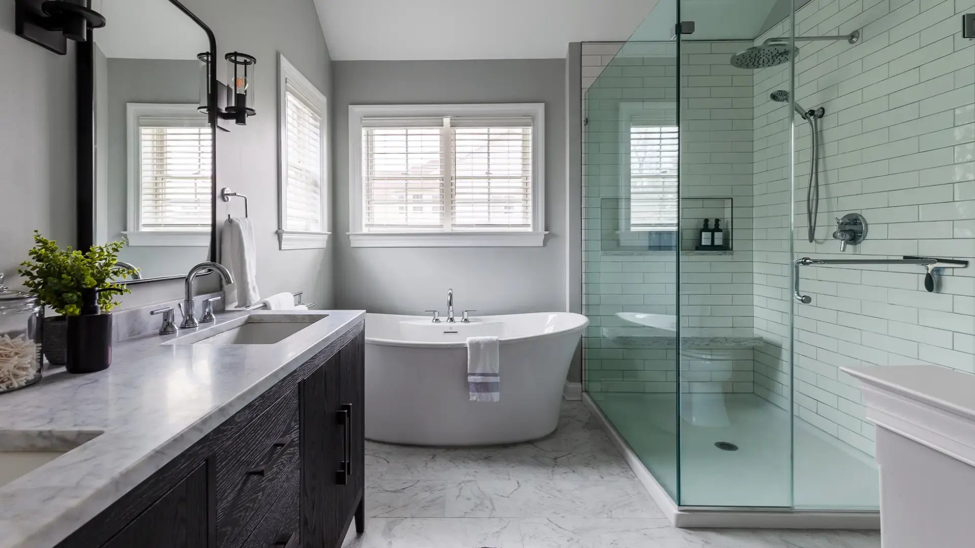 Modern bathroom remodel with white tile floors, dark double vanity with white countertop, large white soaking tub on far wall below a pair of double-hung windows with blinds pulled down, and large glass-enclosed shower on the right with white subway tiles.