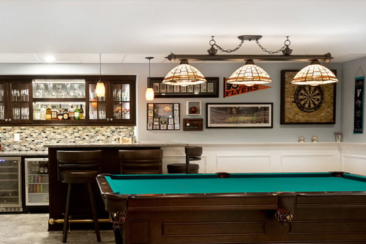 Basement remodel with pool table in foreground, bar area in background with dark cabinets and glass tile backsplash. Wine fridge in lower cabinets.