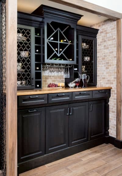 Wellborn Cabinet black cabinetry in a home bar area with light wood countertop, light brick backsplash and wall, and light wood floors.