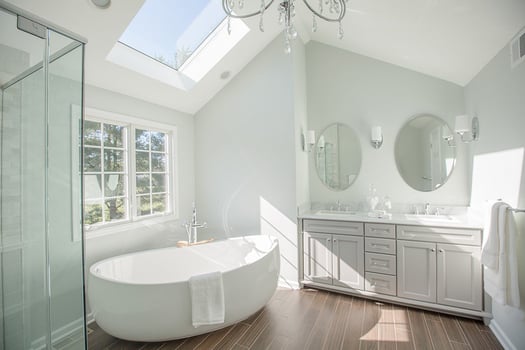 Primary bathroom remodel. Light gray double vanity, skylight over white garden tub, glass shower, wood-look tile flooring.