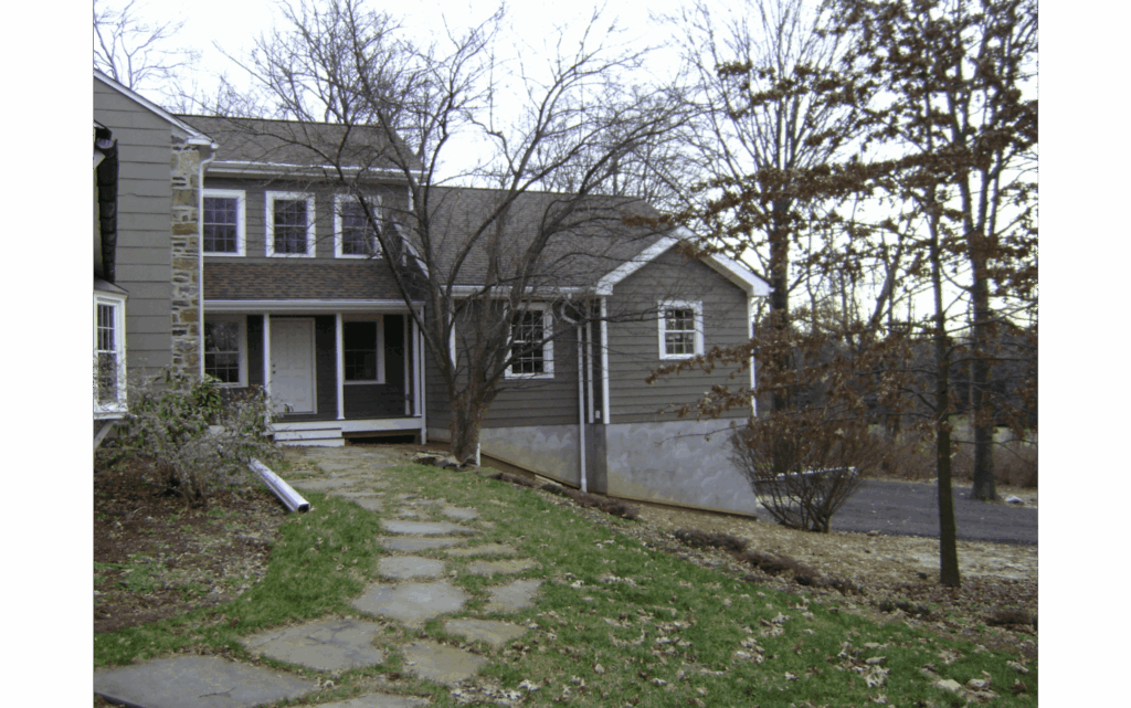 Front of a two-story home with green/gray siding and white trim and small covered front porch, with addition built onto the right side, including a garage underneath. Stone pathway leading to front door. Bare trees surrounding.