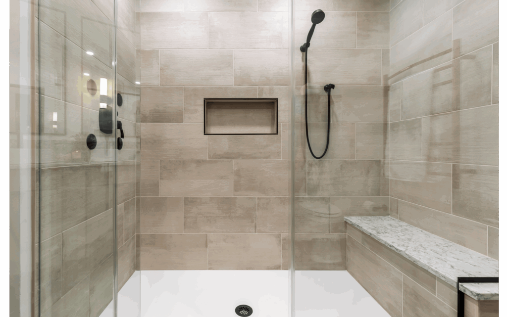 Remodeled glass-enclosed large walk-in shower with white floor tiles, large beige wall tiles, and long bench along right side. Matte black fixtures and detachable showerhead.